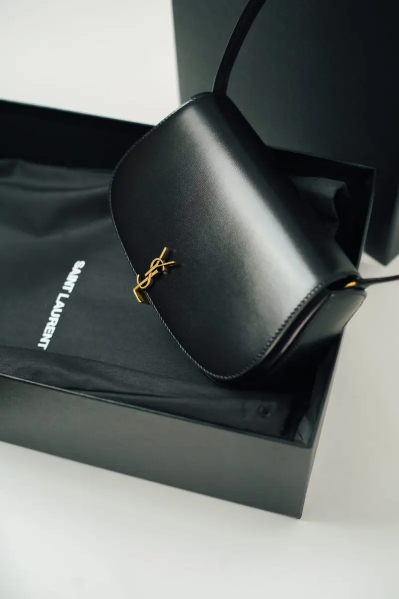A black leather handbag with a gold YSL logo is partially visible inside a black box. A black dust bag with the "SAINT LAURENT" logo is also visible within the box