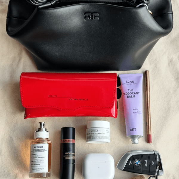 Overhead shot of a black leather handbag with a silver logo closure at the top, placed above a red patent leather wallet with "KHAITE OLIVER PEOPLES" embossed on it. To the right of the wallet is a lavender tube labeled "THE DEODORANT BALM" and a brown lip liner. Below the wallet are several beauty products: a rectangular clear perfume bottle labeled "REPLICA", a black tube labeled "NUDIES MAT", and a small white jar labeled "KIEHL'S". At the bottom, a white wireless earbud case and a car key fob are visible. All items are arranged on a light-colored, textured surface.