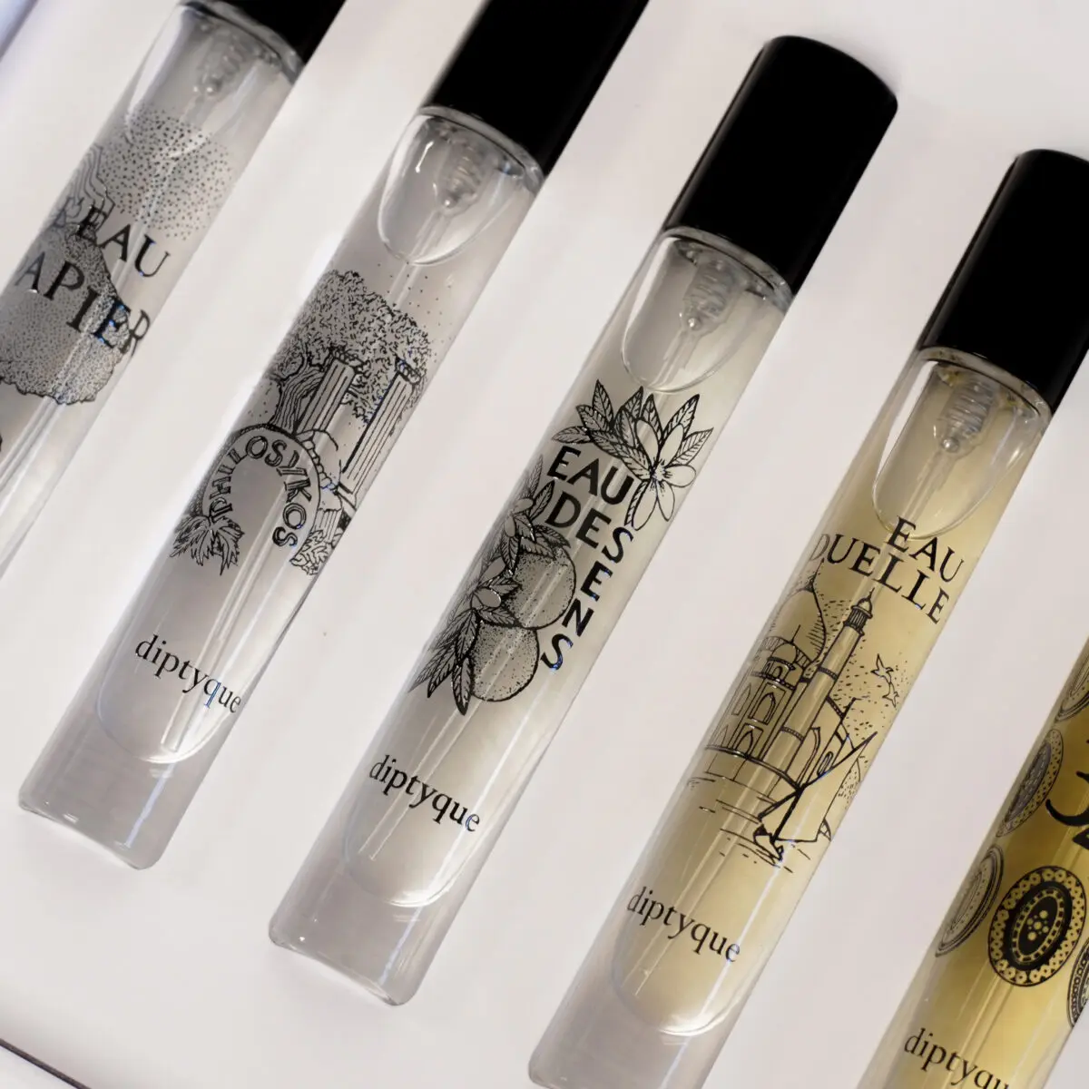 Close-up of five clear glass perfume vials with black caps, each with a different label design and the Diptyque logo