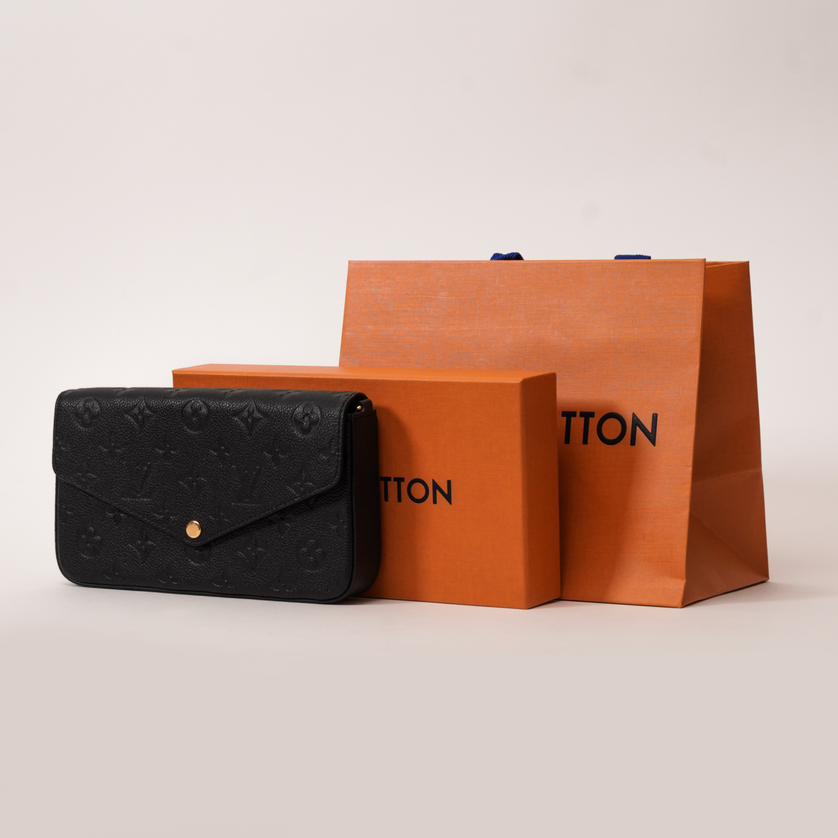 Black Louis Vuitton Felicie Pochette handbag is positioned on the left side of the frame, leaning slightly against an orange Louis Vuitton gift box. Behind the box and partially obscured by it is a larger orange Louis Vuitton shopping bag with blue fabric handles