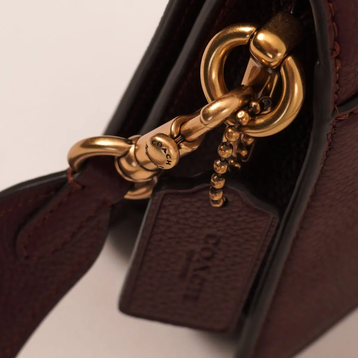 A close-up shot focuses on the gold-toned hardware of a dark brown Coach handbag. A circular ring connects to a lobster clasp engraved with "COACH" in capital letters