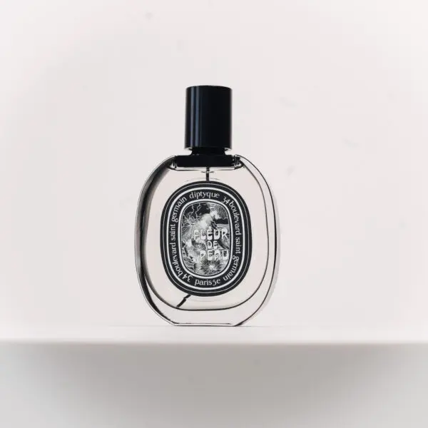 A Diptyque Fleur de Peau perfume bottle in a clear, rounded glass with a black cap, sitting on a white shelf against a white background. The bottle has a white label with black text