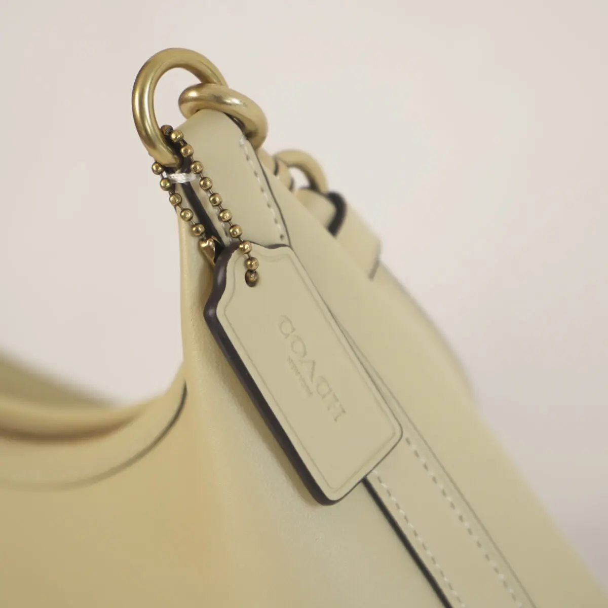 close-up shows a light yellow Coach handbag with visible stitching. A small, rectangular tag with "COACH New York" embossed on it hangs from a brass ring by a small chain