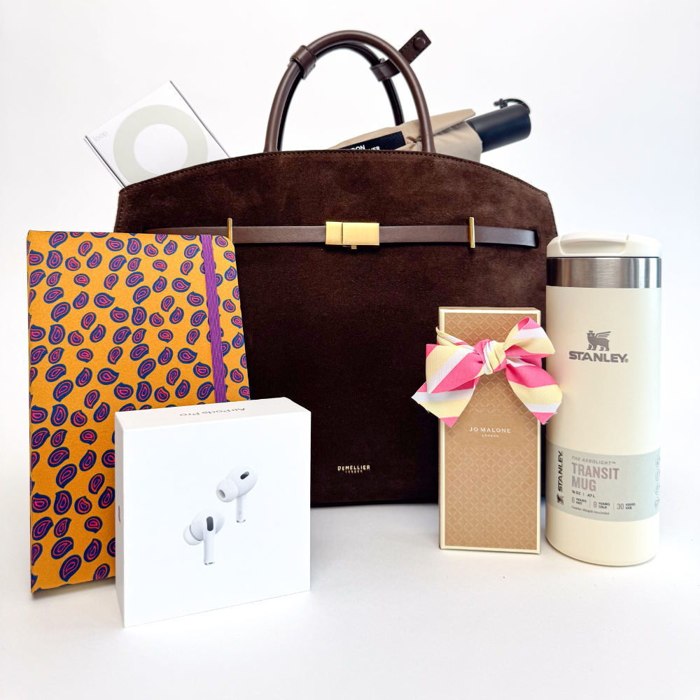 A top-down view of a stylish collection of items, all arranged neatly in front of a dark brown suede bag. From left to right, there is a spiral notebook with an orange and blue paisley pattern, a white box for a pair of Apple AirPods Pro, a small white and blue box for a pair of AirPods (unspecified model), a tall rectangular box for Jo Malone cologne with a striped ribbon bow, and a cream-colored Stanley travel mug with a silver cap.